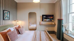 A neatly arranged bedroom with a bed, a large mirror, a TV on the wall, and a window with curtains.