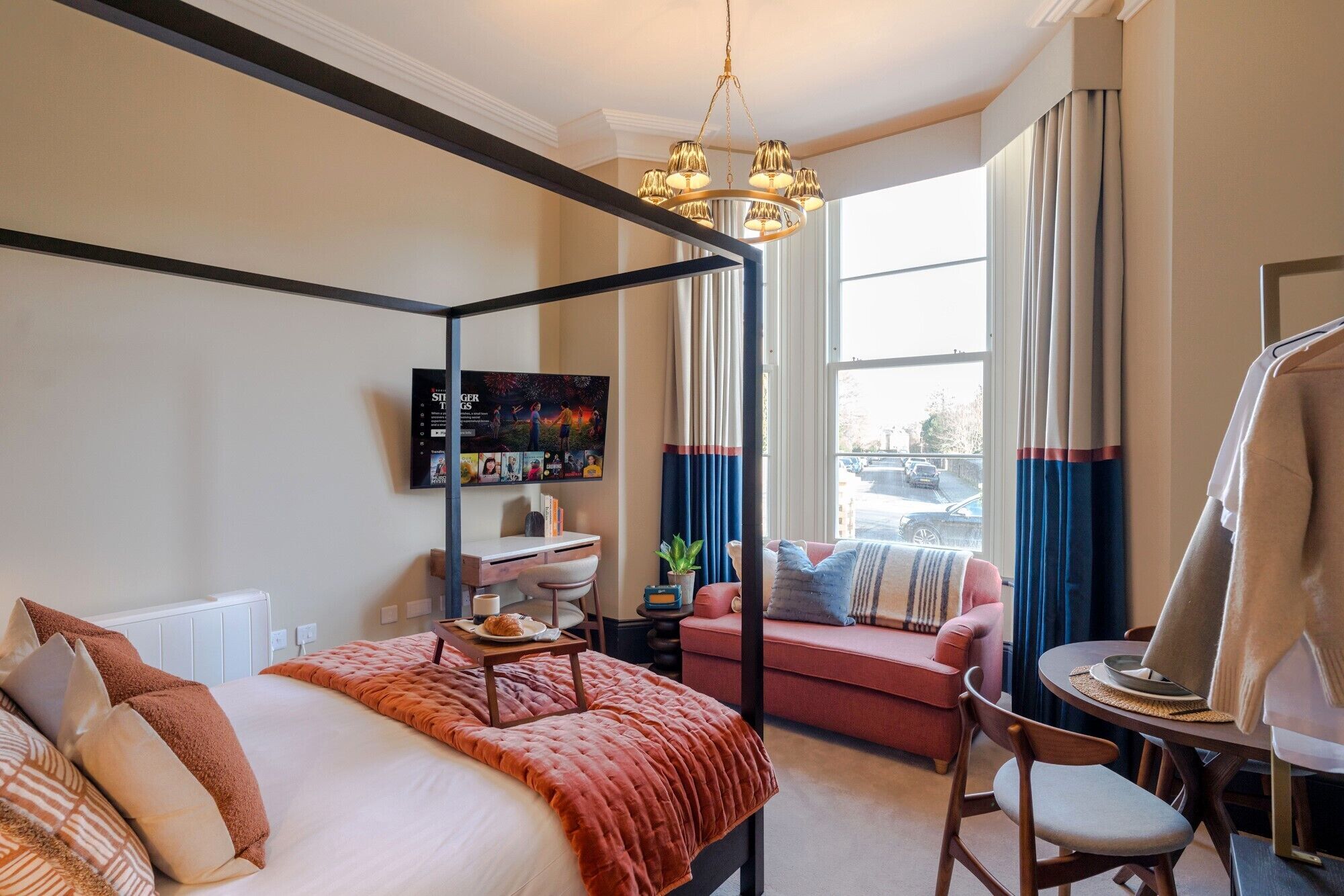 Une chambre à coucher avec un lit à baldaquin, un bureau avec une chaise, une petite table avec une lampe et une grande fenêtre avec des rideaux.