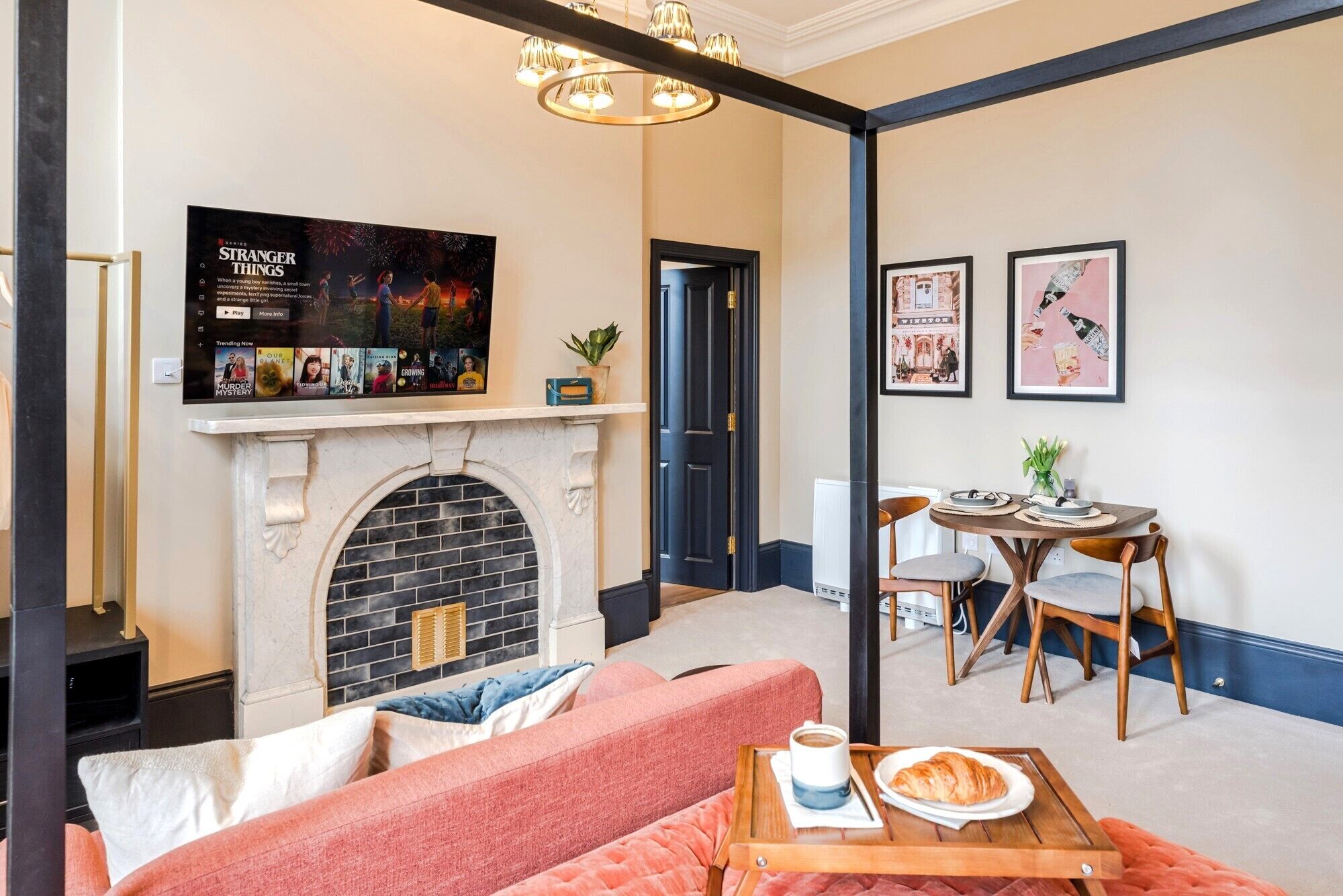 A cozy living room with a fireplace, a sofa, a dining table with chairs, and a TV mounted above the fireplace displaying a movie poster.