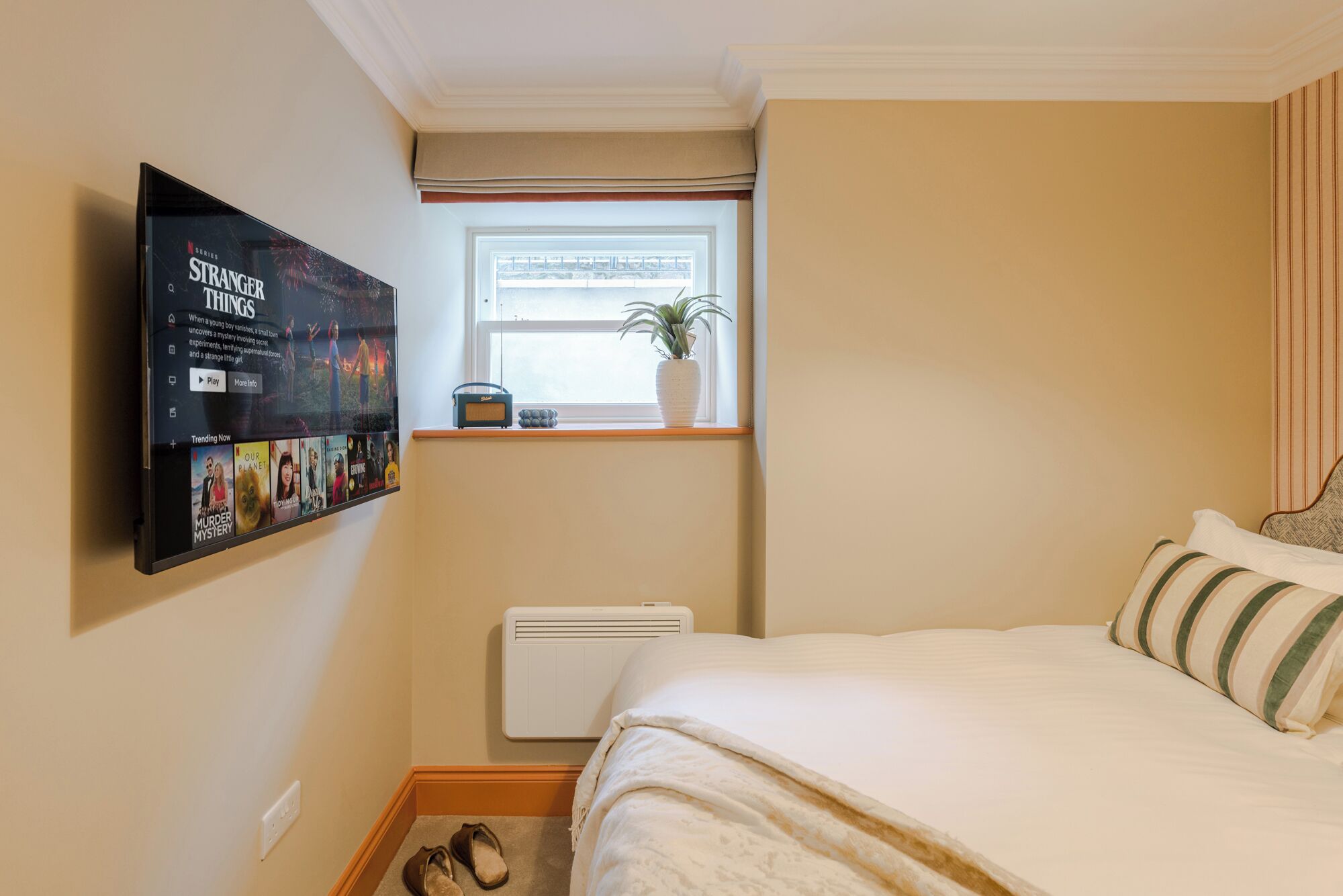 Un dormitorio con cama, ventana con planta, radiador y televisor fijado a la pared mostrando una serie.