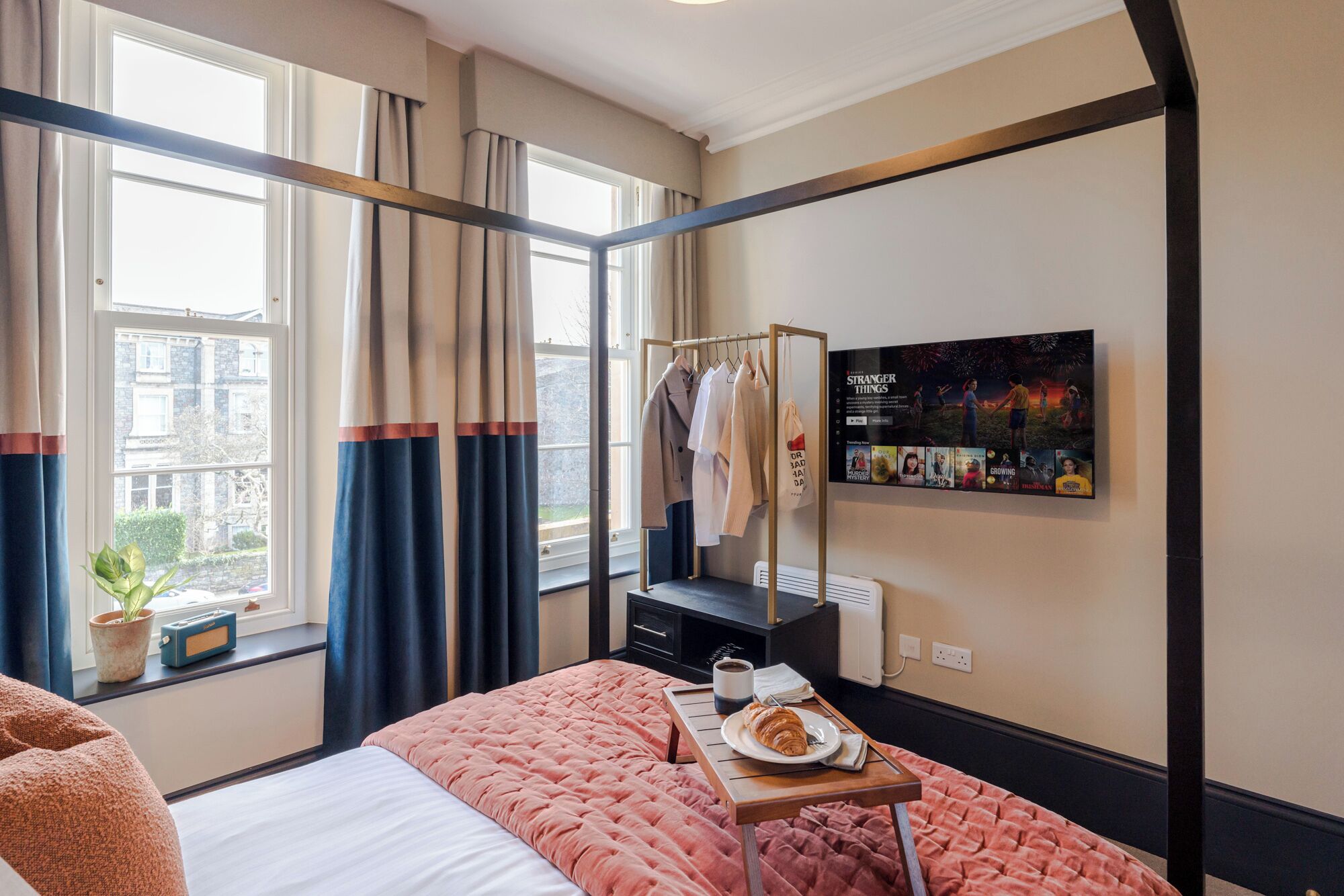 A hotel room with a bed, a bedside table with a croissant and coffee, a coat rack with clothes, a window with curtains, and a wall with a movie poster.