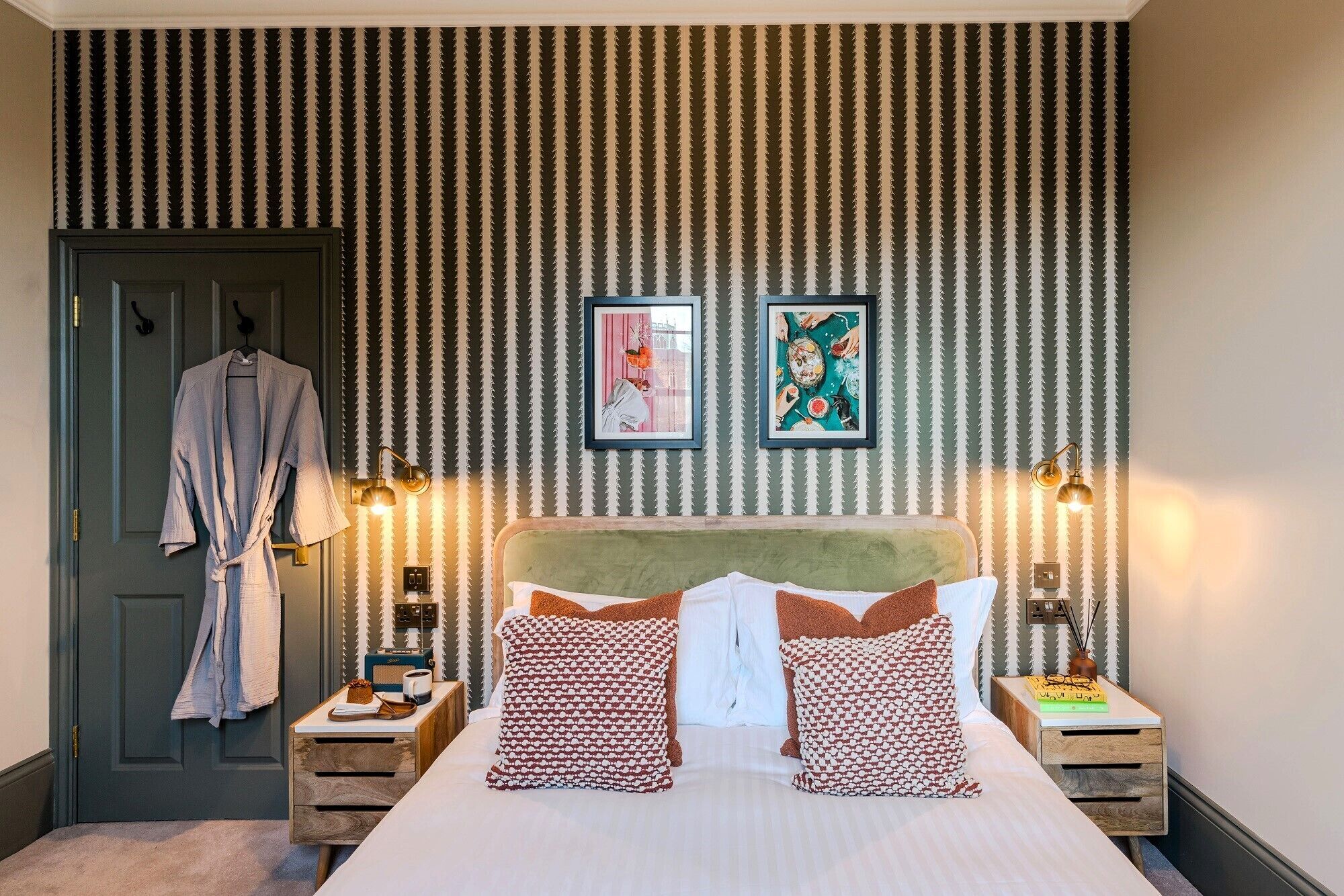 A hotel room with a striped wallpaper, a bed with patterned pillows, two framed artworks, bedside lamps, and a robe hanging on a door.