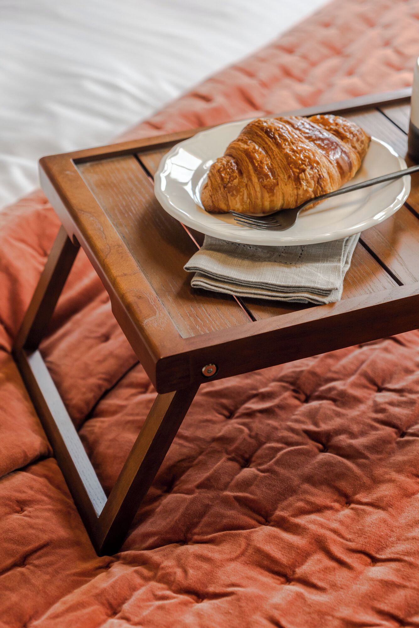 A wooden bedside table with a plate of croissant and a fork on a bed with a red blanket.