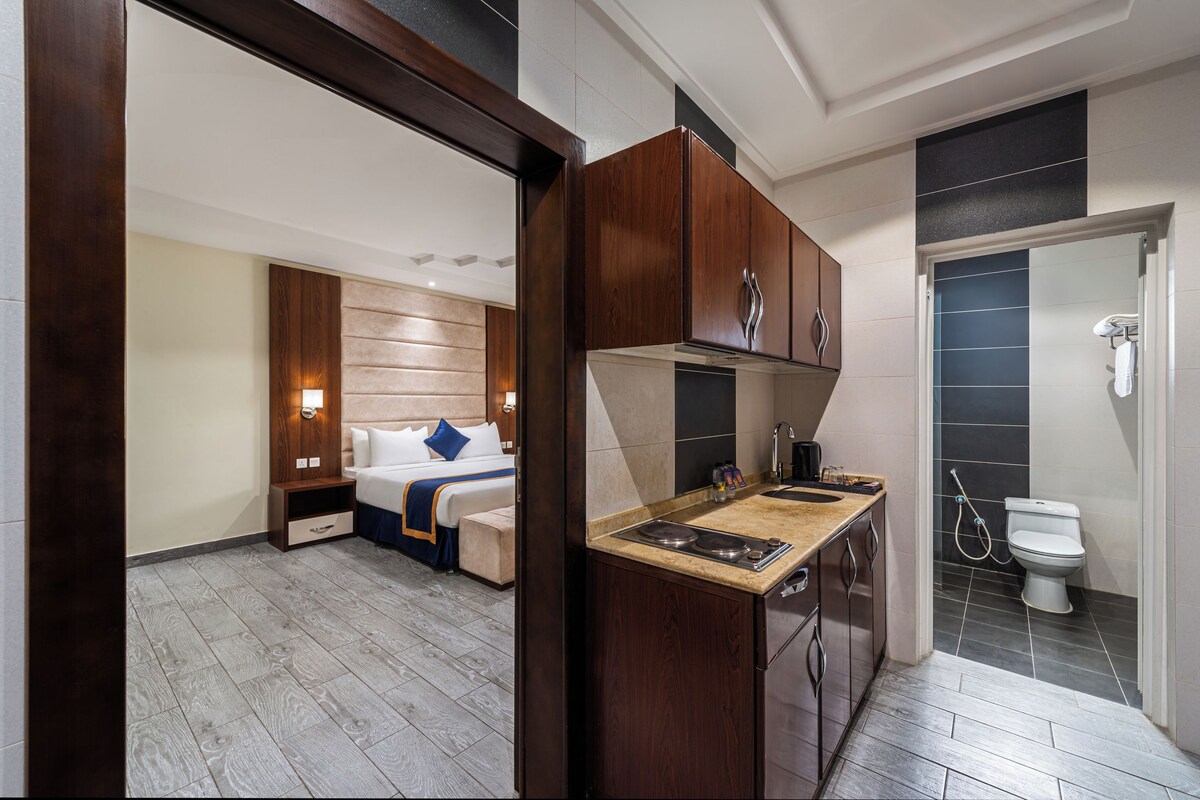 A modern hotel room with a wooden cabinet, a kitchenette, a bed with a headboard, and a bathroom with a toilet and shower.