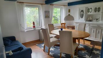 Ein Zimmer mit Esstisch, Stühlen, einer Couch, einem Fenster mit Vorhängen, einem Heizkörper und Wanddekorationen.