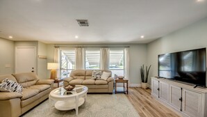 Una sala de estar con un sofá color beige, una mesa redonda para café, un televisor sobre un mueble bajo y un ventanal con cortinas.