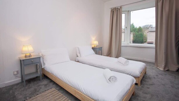 Een slaapkamer met twee eenpersoonsbedden, een raam met gordijnen, een nachtkastje met een lamp en een vloerbedekking.