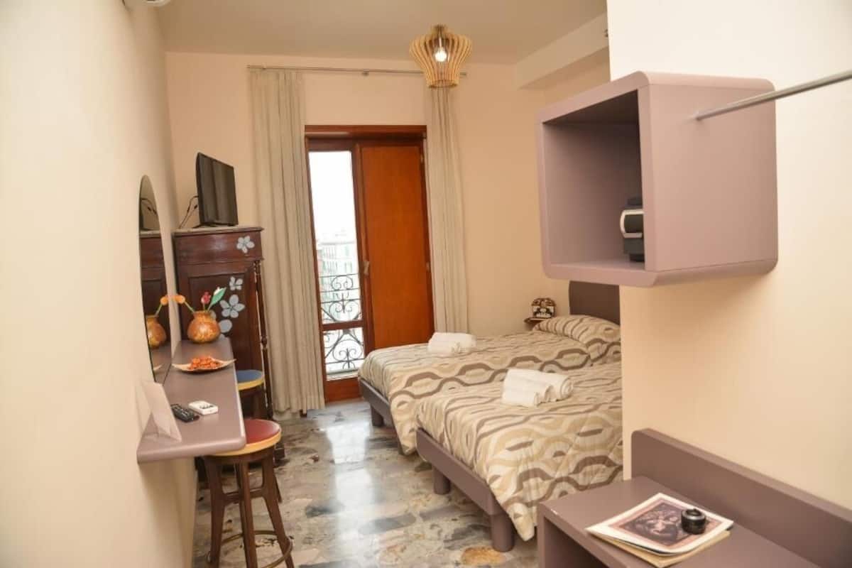A bedroom with a bed, a desk, a chair, a small table, and a shelf.