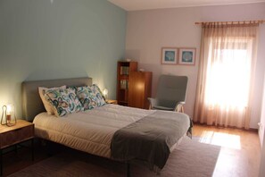 A bedroom with a bed, a chair, a nightstand, a bookshelf, and two wall-mounted pictures. - Seahorse House, T2 Tavira – Santa Luzia (Caminho das Trindades)