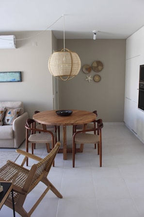 Un comedor con una mesa redonda de madera, sillas de mimbre, una lámpara colgante y una cocina al fondo.