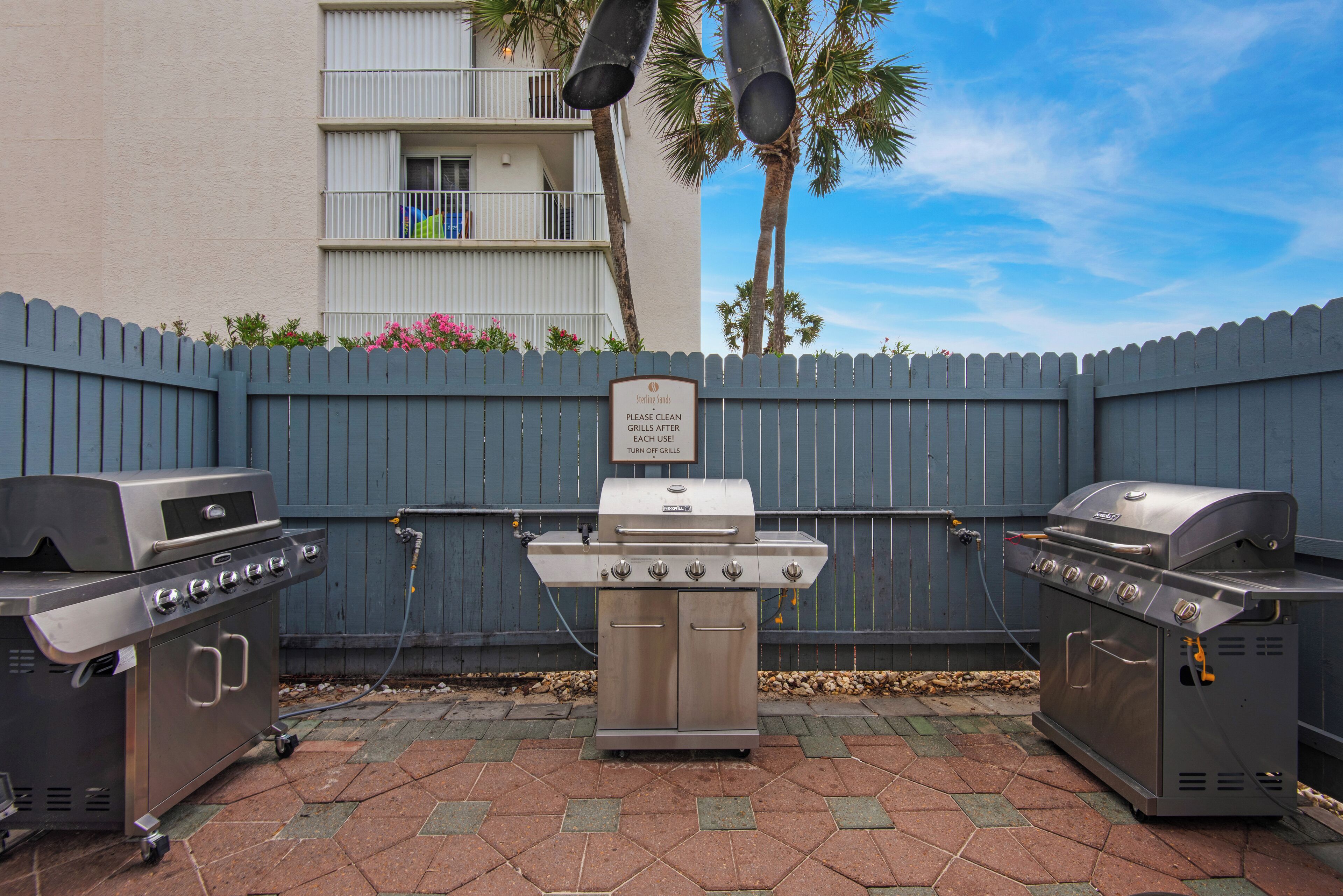 A barbecue setup with two grills and a central grill, set against a blue fence with a sign that reads "Please clean grills after each use."