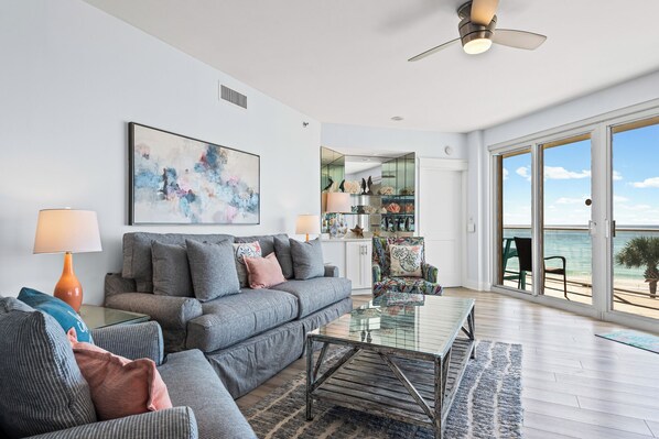 A modern living room with a sofa, coffee table, and a view of the ocean through large sliding glass doors. - Gulf-front 3BR corner unit, updated, balconies, Pool and Hot Tub! (Destin)