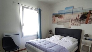 Une chambre à coucher comprenant un lit, une chaise, une table de chevet, une lampe, un ventilateur de plafond et une fenêtre avec des rideaux.