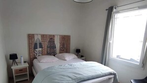 Une chambre avec un lit, une table de chevet, une lampe, une chaise, une fenêtre et un mur décoré d’une fresque représentant une planche de surf.