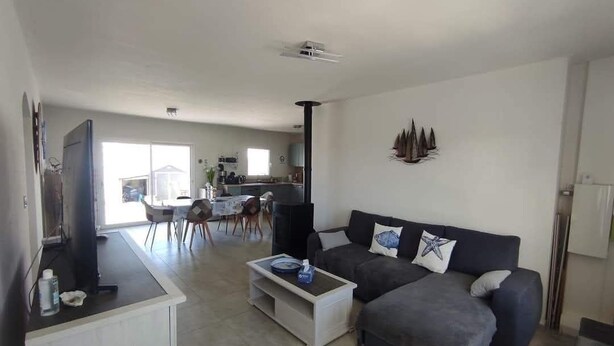 Un salon moderne avec un canapé de couleur foncée, une table basse blanche et un téléviseur à écran plat. On aperçoit, au fond, un coin cuisine avec une table à manger et des chaises.