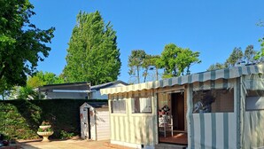 A small, striped-roofed structure with a window and a door, situated in a paved area with a white fence in the foreground. - Mobil home 4 places terrasse couverte à 2 pas de la plage. Animaux non acceptés (Marennes-Hiers-Brouage)