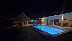 A swimming pool with lounge chairs and a thatched-roof structure, surrounded by a well-lit patio area.
