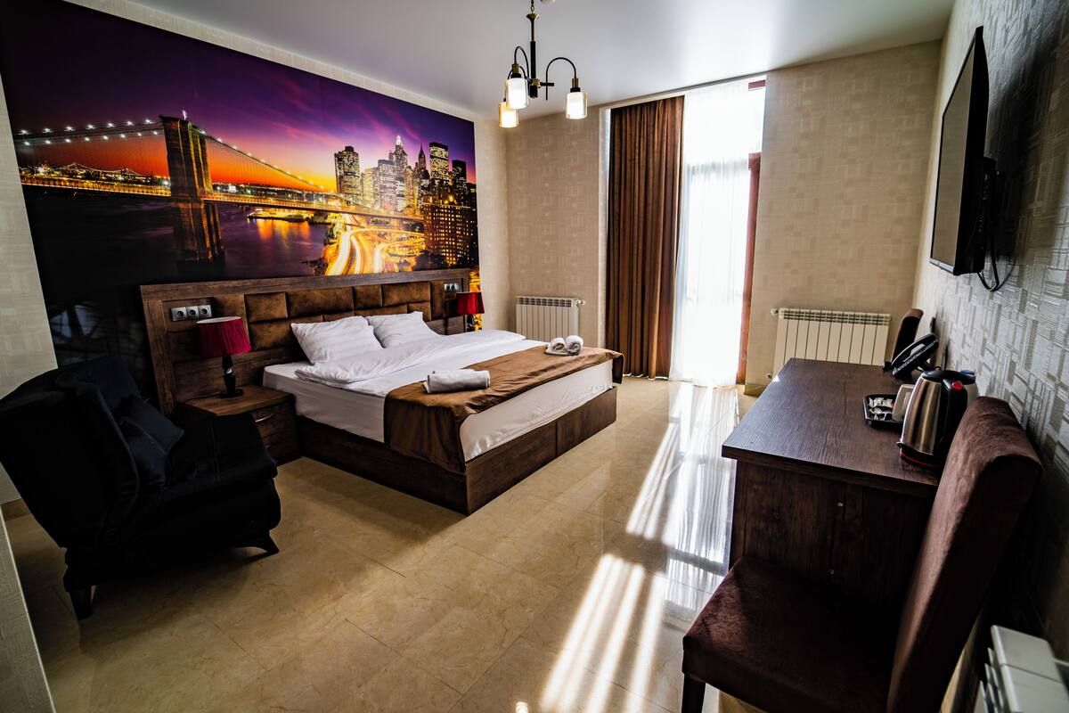 A hotel room with a large bed, a desk, a chair, a radiator, and a cityscape mural on the wall.