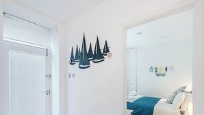 A bedroom with a bed, a nightstand, a wall-mounted shelf with decorative items, and a door with blinds. - 2 bedroom accommodation in Looe (Looe)