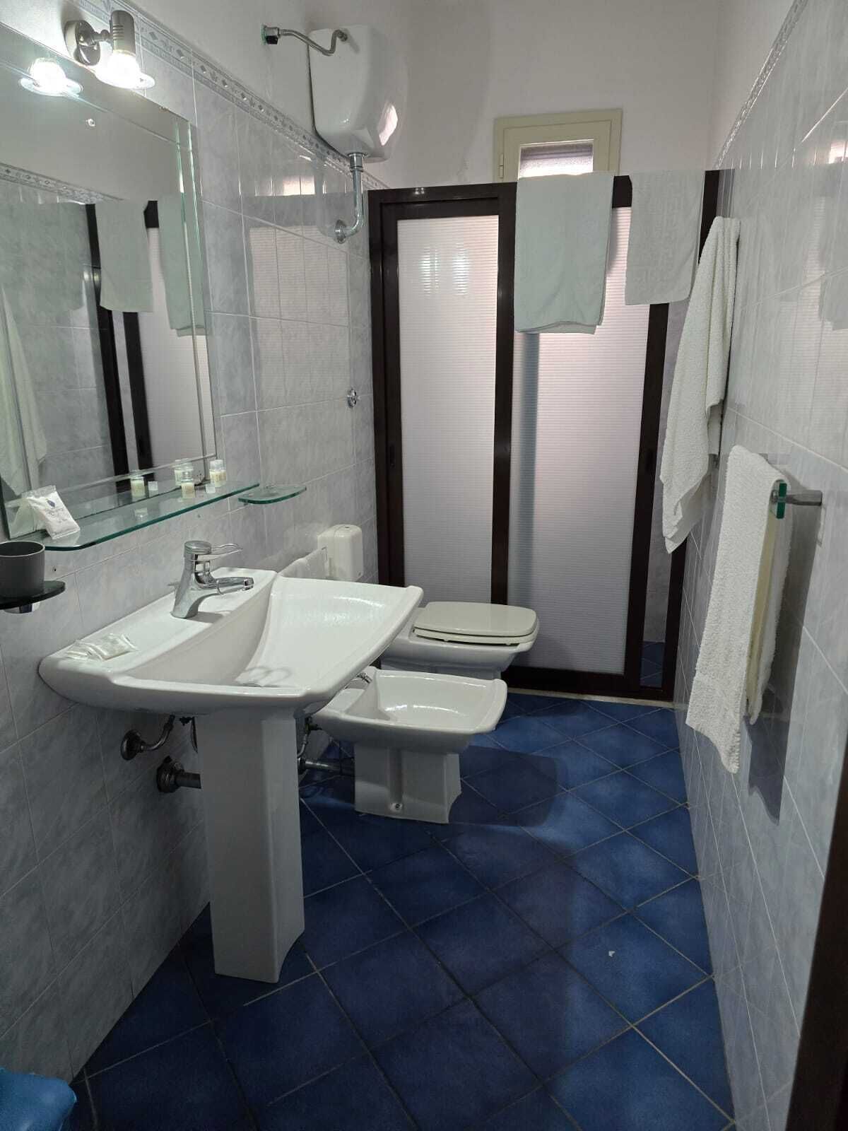 Un baño con un lavamanos blanco, dos inodoros, un estante de vidrio y un espejo.