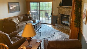 A cozy living room with a fireplace, a ceiling fan, and a large window overlooking a deck. - 2BR Village At Indian Point 1306 near SDC - Free Tickets Daily up to $500 (Branson)