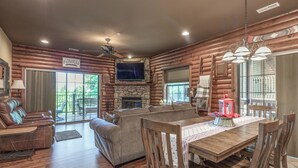 L’intérieur d’une cabane en rondins présente une cheminée en pierre, un téléviseur fixé au-dessus, un coin repas avec des chaises et une table en bois, ainsi qu’un ventilateur de plafond.
