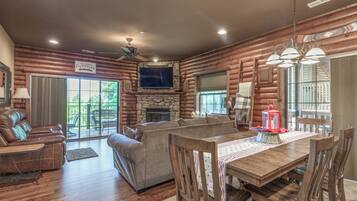 A log cabin interior with a stone fireplace, a television mounted above it, a dining area with wooden chairs and a table, and a ceiling fan.