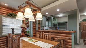 Interior de una cabaña de troncos con mesa de comedor, sillas de madera, zona de bar y cocina con armarios verdes.