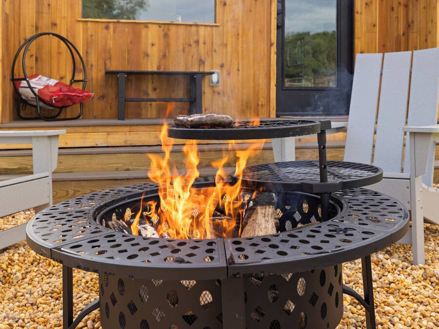 Un barbecue extérieur en métal, avec des flammes, entouré de gravier, avec des chaises longues en bois et un banc à l’arrière-plan.