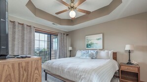 Une chambre à coucher comprenant un lit, un bureau, une table de chevet, un ventilateur de plafond et une fenêtre avec des rideaux.
