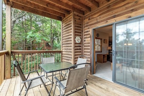 A wooden deck with a table and chairs, a glass door leading inside, and a view of the surrounding forest.