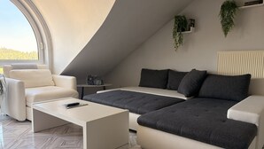 Un salon moderne de style industriel, avec un canapé d’angle blanc, une table basse blanche et une petite table d’appoint. Une grande fenêtre offre une vue sur l’extérieur.