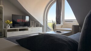 Un salon moderne avec une grande télévision affichant le logo « NETFLIX », un canapé, une table basse et une vue dégagée sur l’extérieur grâce à une grande fenêtre.
