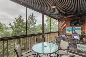 Un espace extérieur abrité, doté d’une table en verre et de chaises en osier, d’un mur en bois orné d’un tableau, et offrant une vue sur des arbres et une étendue d’eau.