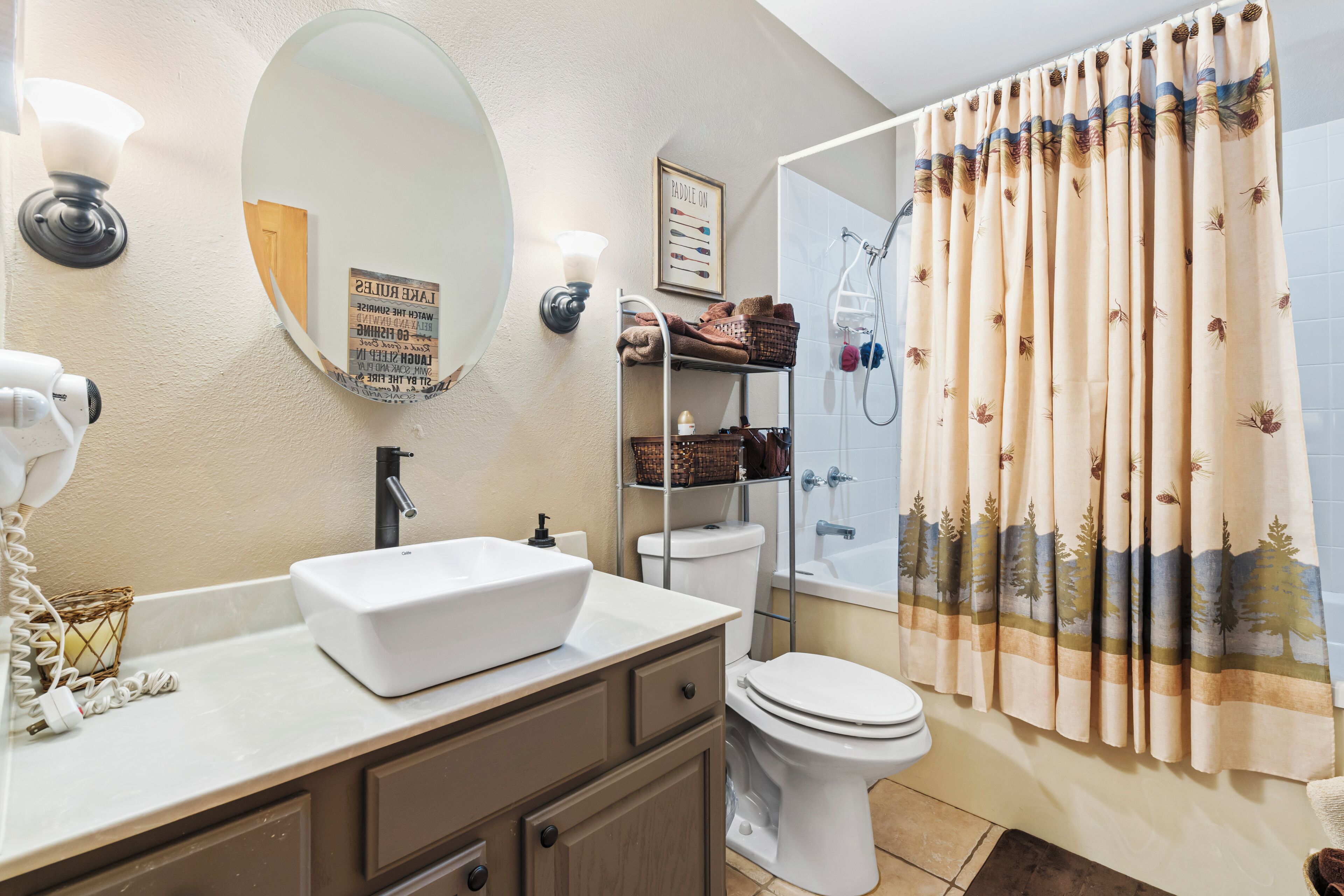 Une salle de bain avec un lavabo blanc, un miroir, un porte-serviettes, des toilettes et un rideau de douche orné d’un motif naturel.
