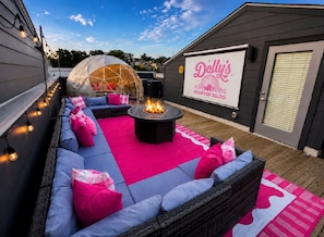 An outdoor seating area with a fire pit, a covered structure, and a large screen displaying 'Dally's Rooftop Igloo'.