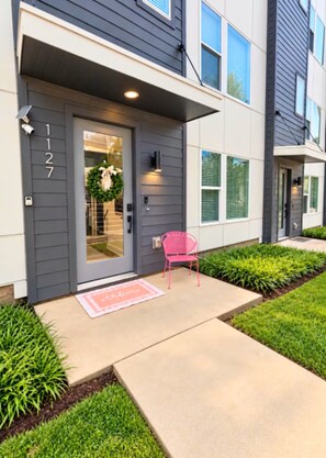 A modern two-story residential building with a grey facade, a front door with a number, a small porch with a pink chair, and a welcome mat.
