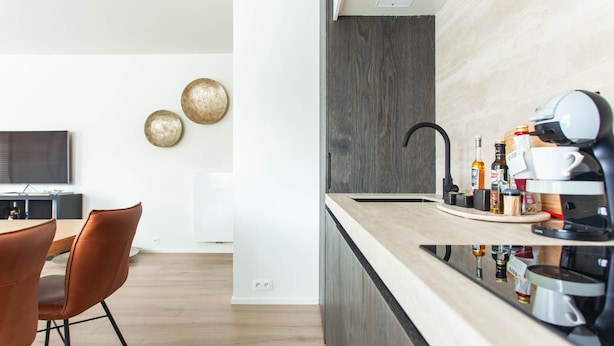 A modern kitchen with a built-in coffee station, a dining area with a wooden table and chairs, and a TV in the background.