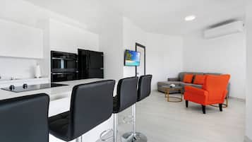 A modern kitchen with a dining area, a grey sofa with orange cushions, and a flat-screen TV on the wall.