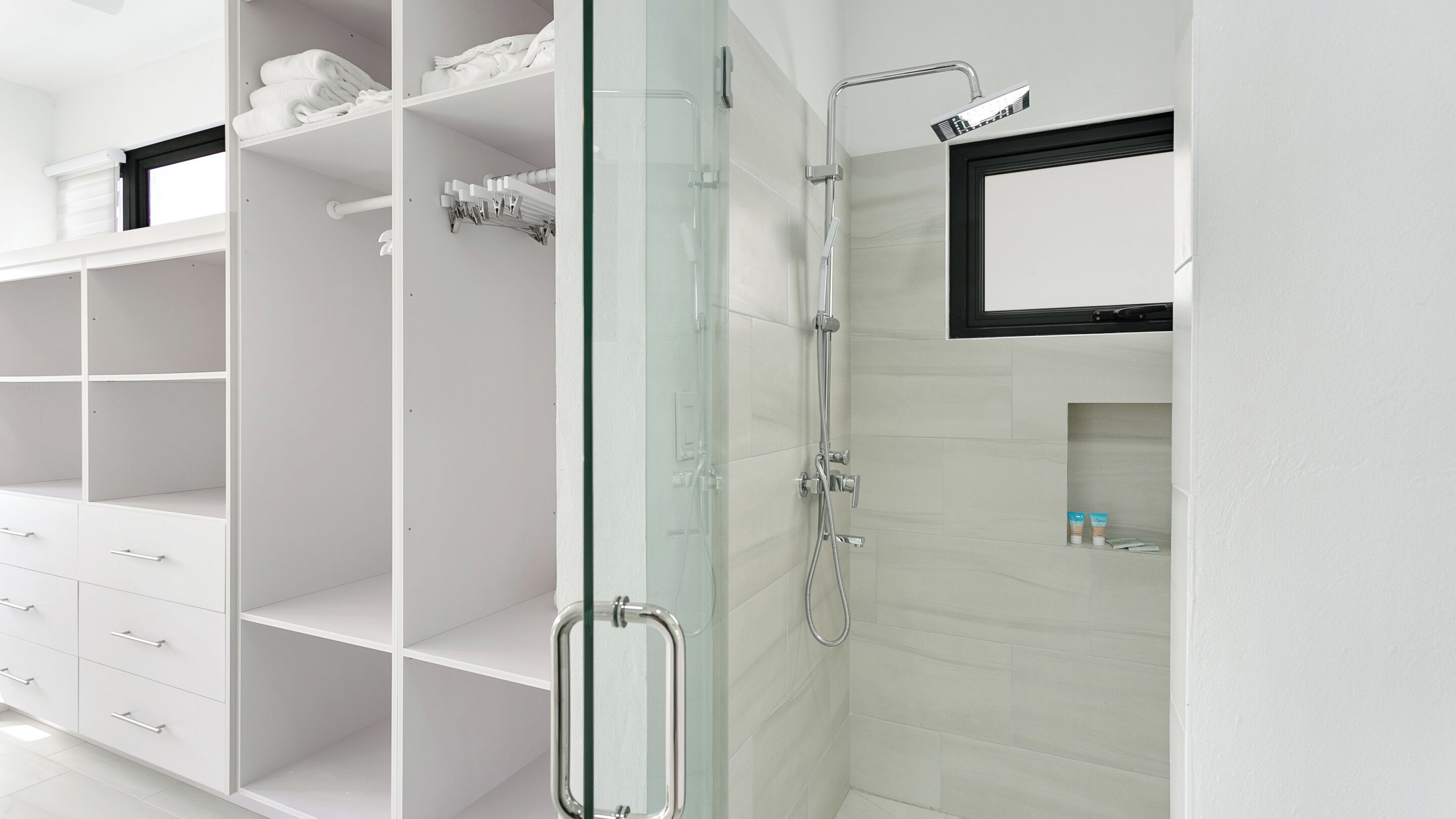Un baño moderno con cabina de ducha de vidrio, tocador blanco con cajones y una pequeña repisa con artículos de aseo.