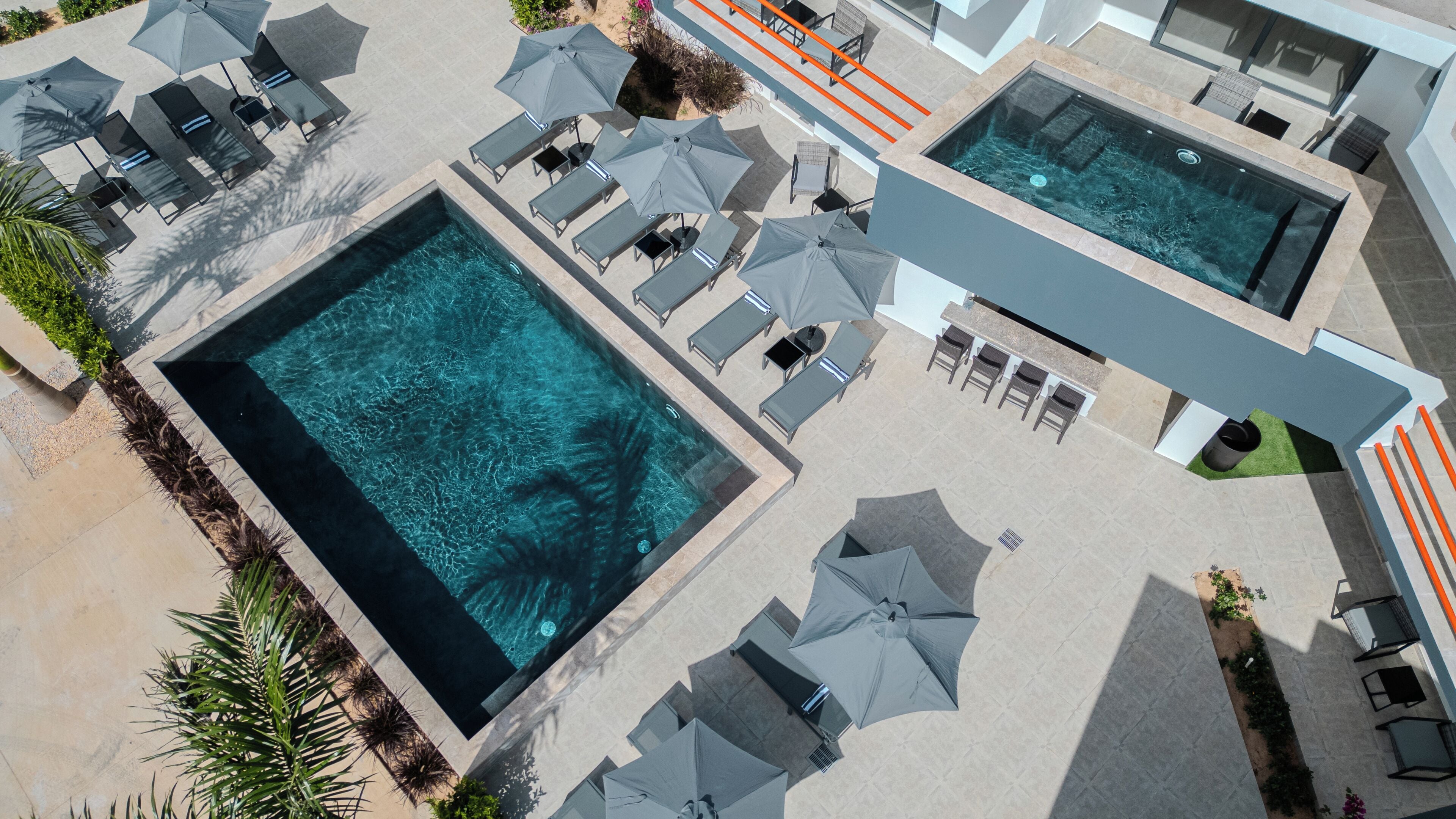 Vista aérea de un hotel moderno con dos piscinas rectangulares grandes, varias zonas de descanso con sombrillas y una zona de bar.