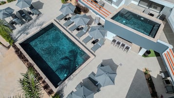 Vista aérea de un hotel moderno con dos piscinas rectangulares grandes, varias zonas de descanso con sombrillas y una zona de bar.