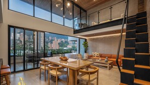 A spacious living area with a large wooden dining table, comfortable seating, a staircase, and a city view through large windows. - Exclusivity in Poblado/Jacuzzi-Morph 1601 (Medellín)