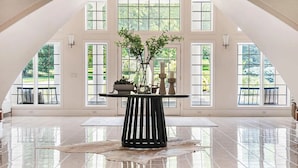 A spacious room with a high ceiling and large windows, a central table with a vase of greenery, and a polished floor.