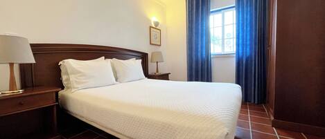 A neatly made bed in a hotel room with a wooden headboard, a nightstand with a lamp, and a window with blue curtains.