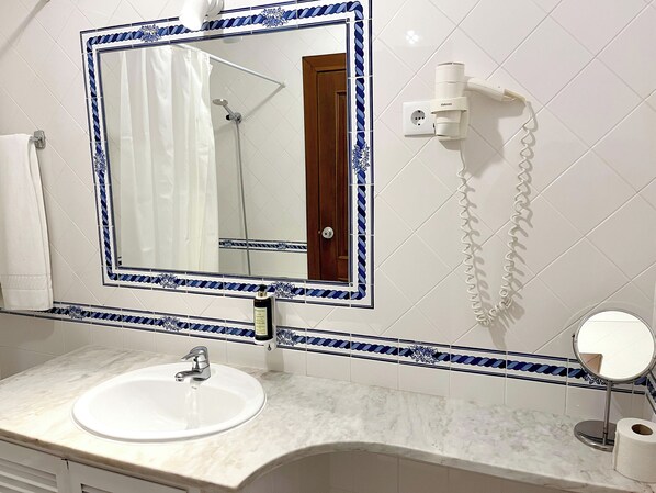 A bathroom with a white sink, a mirror with a decorative frame, a shower, and a toilet paper holder. - Pátios da Vila – Duna Parque Resorts & Hotels (Odemira)