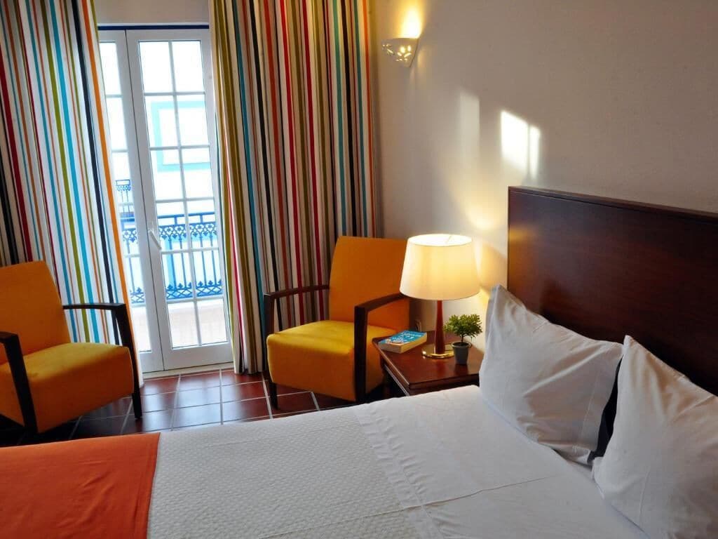 A hotel room with a bed, two chairs, a nightstand with a lamp, and a view of a balcony with colorful curtains.