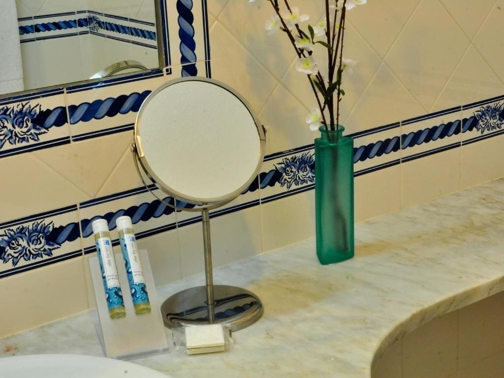 Ένα μπάνιο με έναν στρογγυλό καθρέφτη τοποθετημένο σε βάση, ένα πράσινο βάζο με λευκά λουλούδια και δύο μπουκάλια λοσιόν πάνω στον πάγκο.