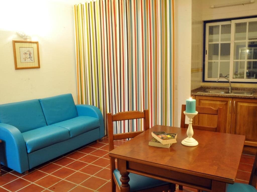 A living room with a blue sofa, a wooden dining table with chairs, a striped curtain, and a kitchen area with a window.