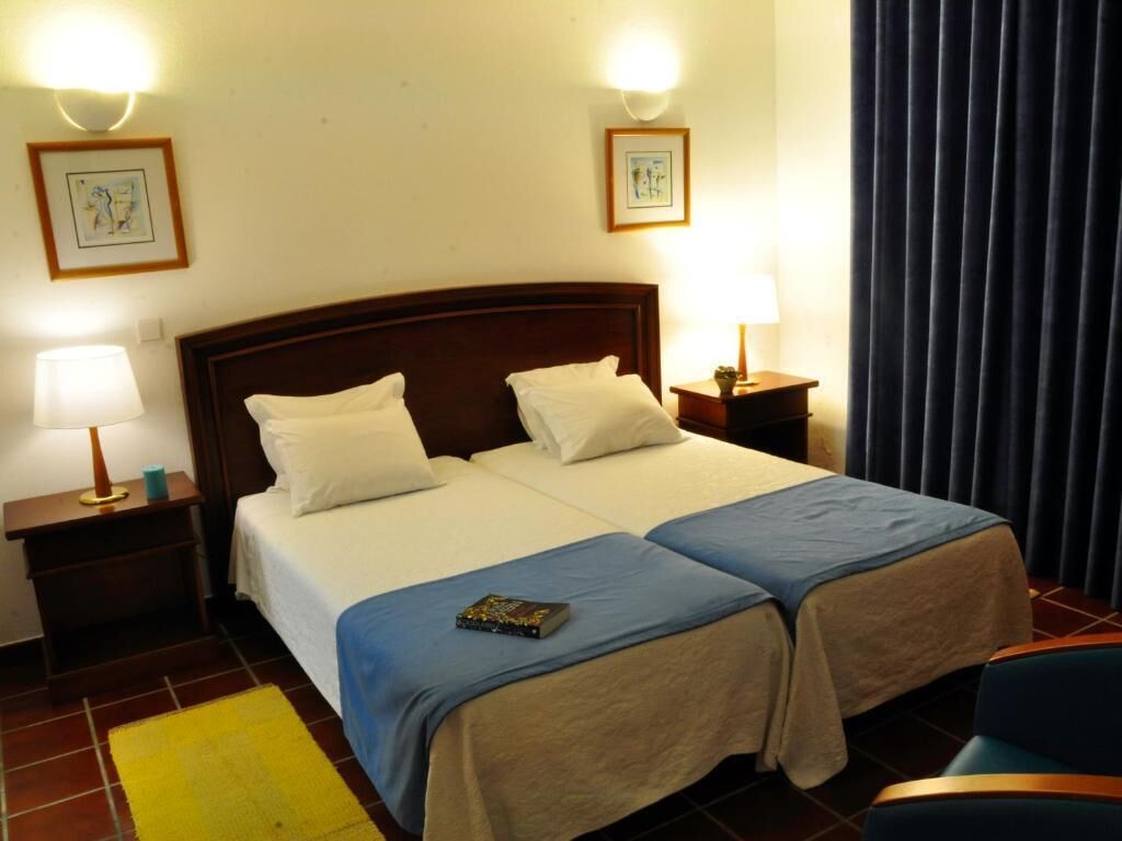 Una cama doble con cabecera de madera, dos mesitas de noche con lámparas, una alfombra amarilla y un cuadro enmarcado en la pared.